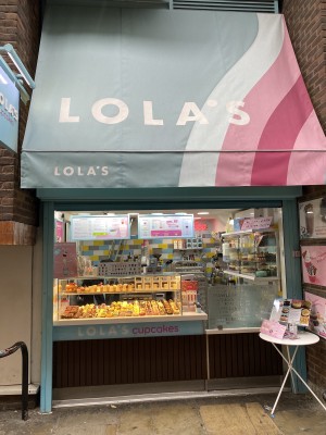 Directly facing Lola’s Cupcakes from the footpath. On left hand side is the glass cabinet with cupcakes. In the middle is the service counter. On the right hand side is a door to the kitchen. In front is a table with menus on top.