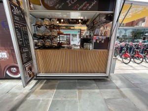 From street, kiosk counter ahead. Paths to outdoor seating area either side of kiosk.