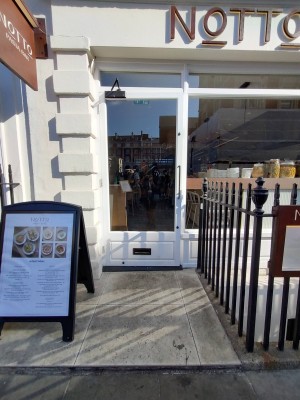 Glass door with white frame. Silver vertical handle on right. Push to open. A-frame board on left. Black metal gates on right.