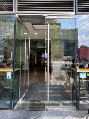 Double glass doors in glass frame. Push vertical metal handle at centre to open. Doorbell for assistance on left of door.