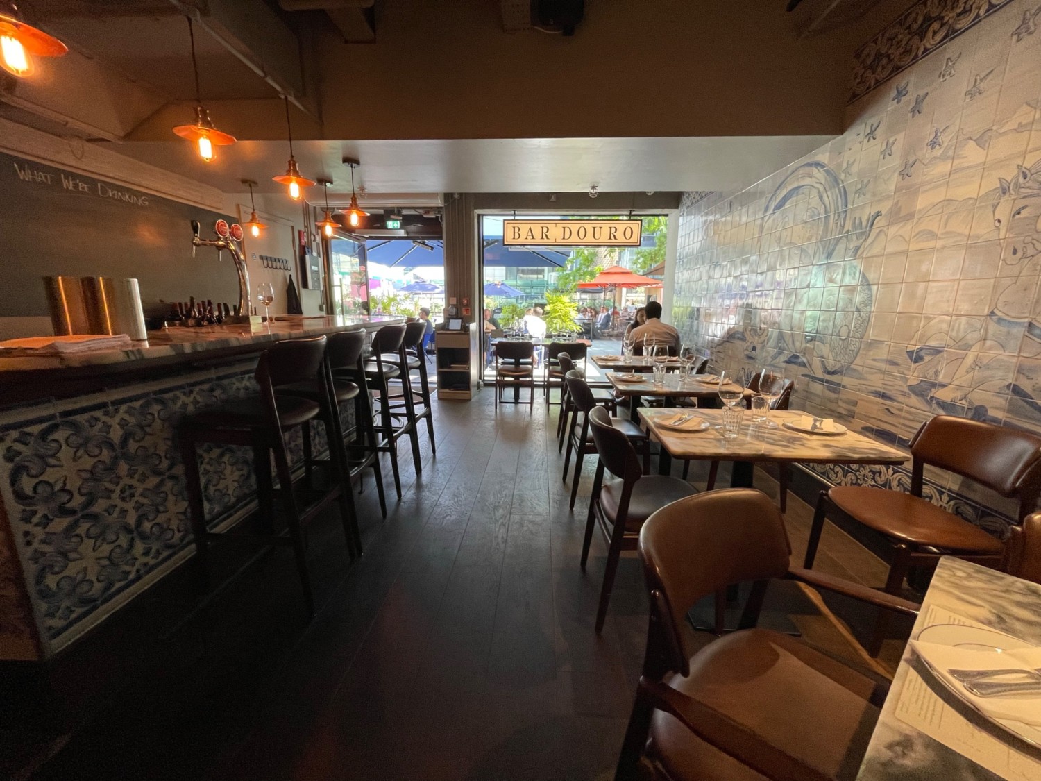 From refrigerator, facing entrance. Tables and chairs along right wall. Path ahead. Bar along left. Glass wall at far back.