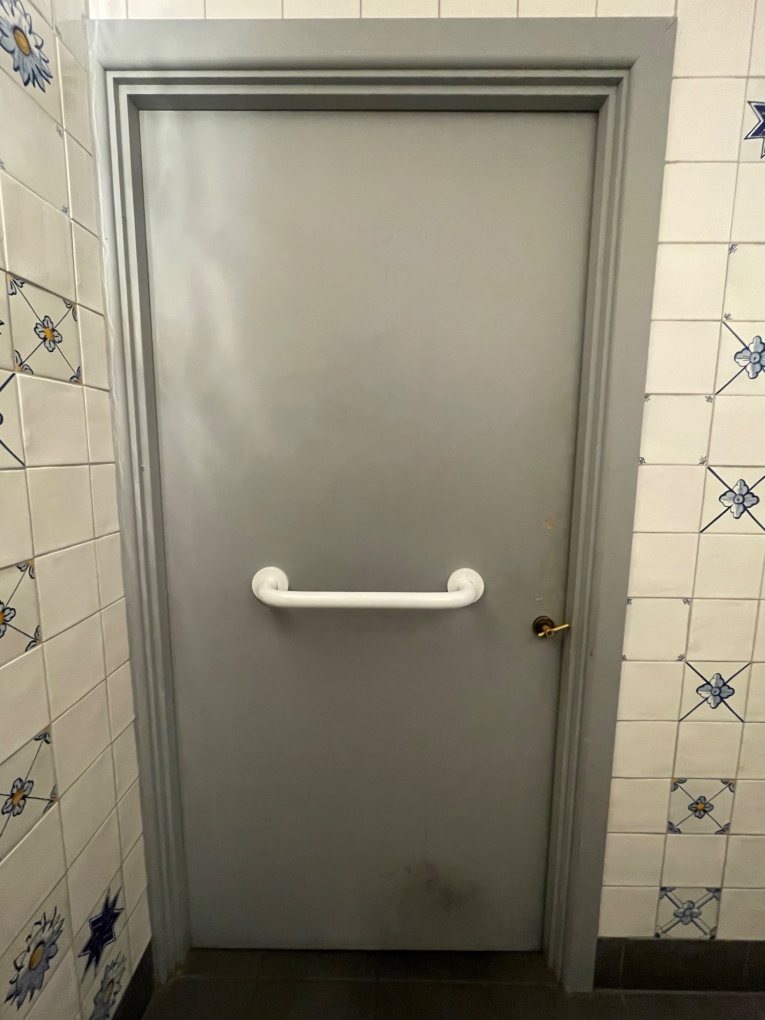 From sink, facing back of toilet door. Grey door with white metal grab rail along centre. Push right side to open.