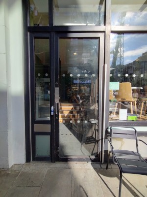 Glass doors with brown frame. Silver vertical handle on left. Push to open. Doorbell immediately on left of handle.