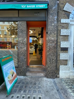 Facing entrance. Orange wood-framed door ahead. Two steps up to doorway. Push silver handle on left to open. Marble and glass walls either side.