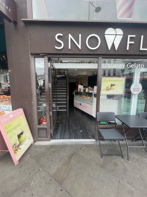 Glass door with dark brown frame. Push to open. Sandwich board to left of door. Tables and chairs to right.