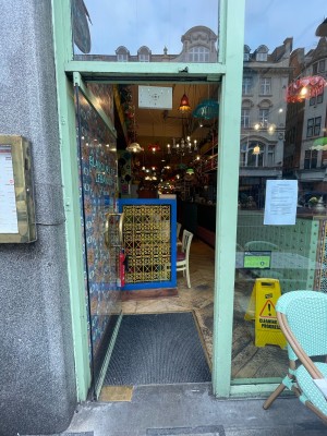 From pavement, facing straight. Glass door with green frame ahead. Gold metal handle to right. Push to open. Blue and yellow partition ahead of door.