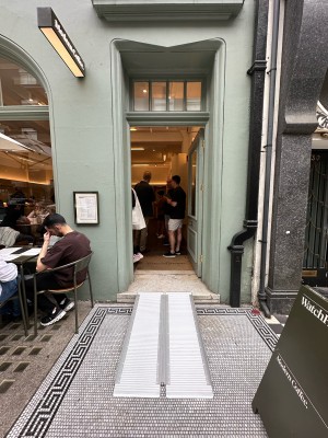 Green and glass panelled door. Gold handle on left, push to open. Ramp laid out leading up to door.