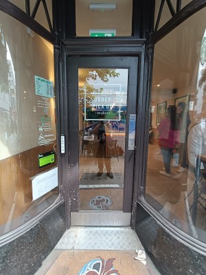 Single glass door with brown wooden frame. Silver metal pad right. Push to open. Doorbell on left doorframe.