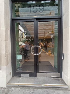 Glass doors with black frame. Silver curved handle in middle. Push to open. Doorbell on right wall.