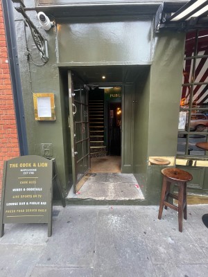 From pavement, facing straight. Step ahead. Entrance door after step. Sandwich board to left.