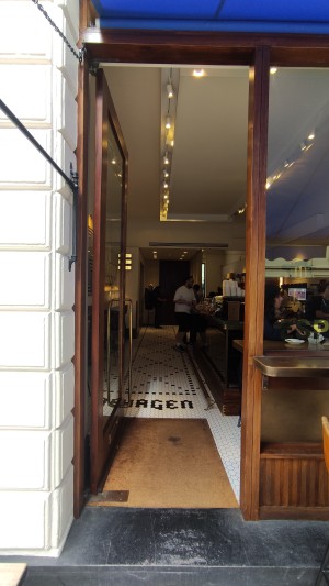 Single glass door with brown wooden frame. Push from right via vertical brown metal handle to open.