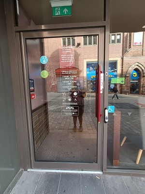 From entrance facing straight. Grey framed glass door ahead. Red pull metal handle at right. Doorbell assistance sensor on right.