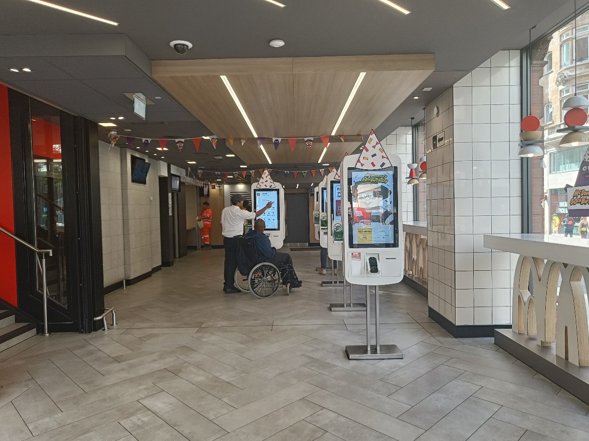 From entrance, looking right. High tables along right. Pillars in between. Ordering screens spread ahead. Counter at back left. Path in between.
