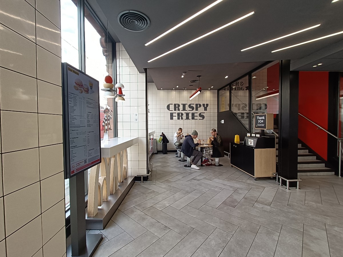 From entrance, looking left. High tables along left. Pillar in middle of tables. Table and stools at back. Bin on right. Stairs at back right.