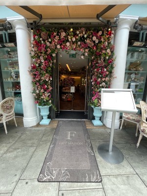From sidewalk facing venue. Menu sign on right. Path with doormat leading to entrance ahead. Large plants to left and right of entrance.