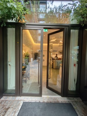 Glass door with brown frame. Push silver vertical handles in centre to open. White curtains after door, pull from centre to open. Doormat before door.