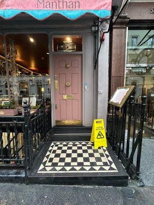 From pavement, facing straight. Opening ahead with step up at foot of it. Black railings along left and right. Menu and A-frame on right.