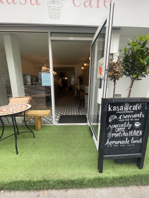 From pavement, facing straight. Step ahead before entrance. Glass door with white frame after step. White handle to left. Pull to open.