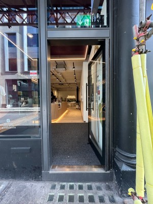 Single glass door with black metal frame and vertical black metal handle along left. Push to open.