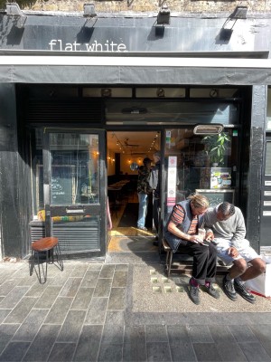 Black wood-panelled glass door ahead. Outdoor seating on left and right. Glass walls either side. Pull silver handle on right side to open.