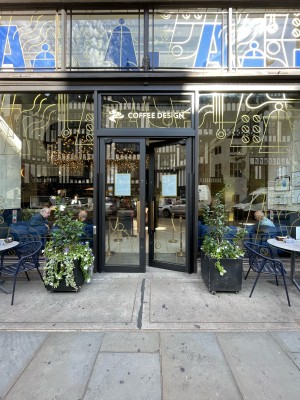 Glass doors with black frames ahead. Gold vertical handles in centre, push to open. Planters to left and right of doors.