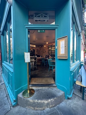 Dark blue and glass panelled door. Push gold pad on right to open. Gold standing sign to left of door on top of step.