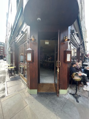 Single glass door in brown wooden frame set back from pavement. Pull gold metal handle from left to open.