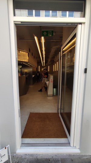 Glass door with white frames ahead. Push to left via vertical metal handle to open.