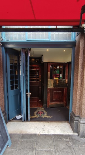 From sidewalk facing venue. Glass sliding door with blue frames ahead. Slide to left to open via brass door handle.