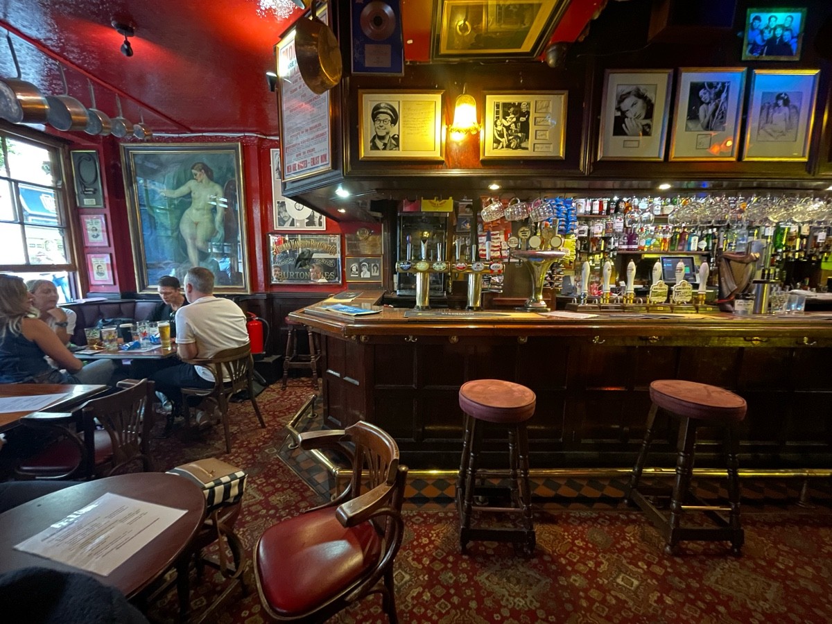 From entrance, facing left. Table and chairs ahead. Half booth and chairs along left wall. Bar with stools along back wall.