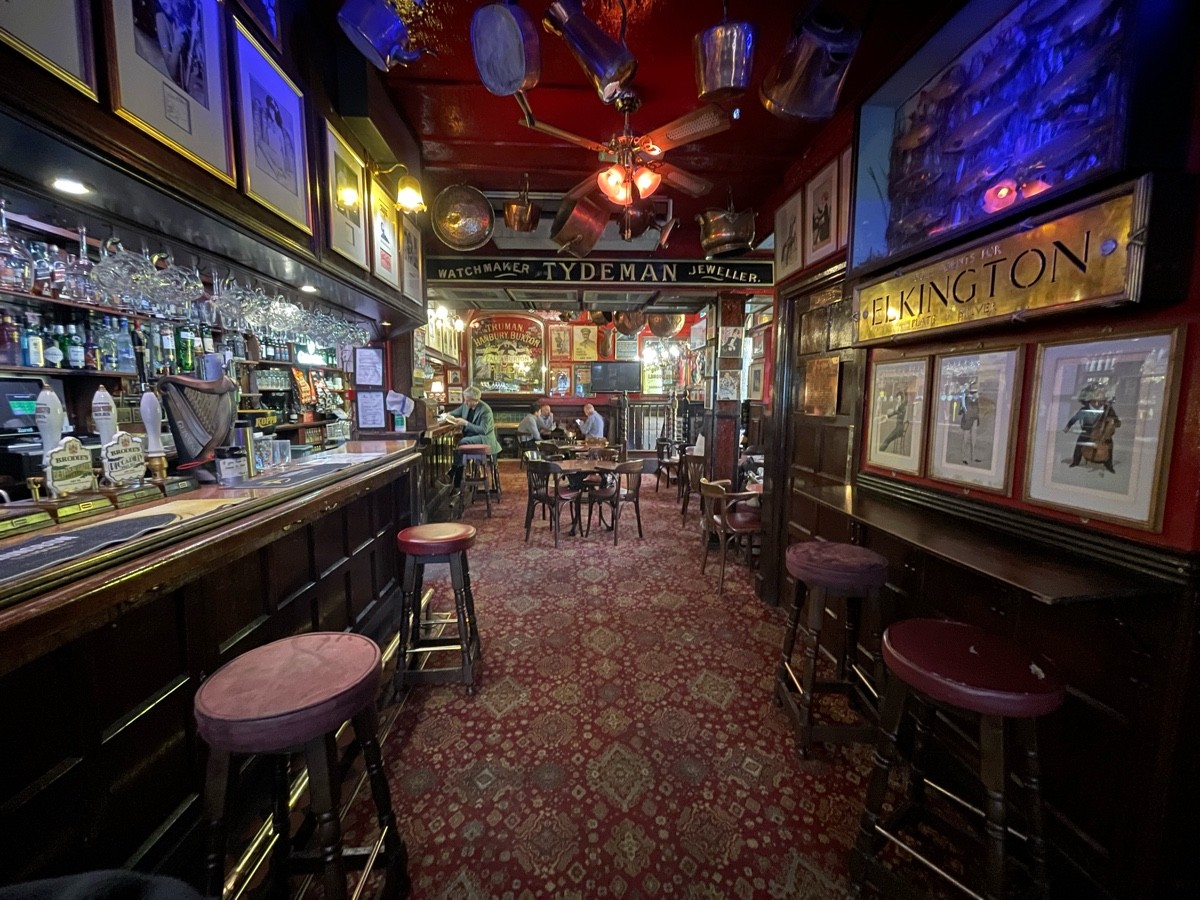 From bar, facing right. Tables and chairs at end of path. Bar with stools along left. High tables and stools along right. Bathroom door beyond it.