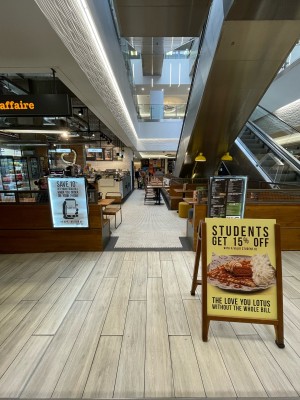 From inside shopping centre facing venue. Sandwich board on right. Half-height wooden dividers ahead back left and right. Open entrance in between.