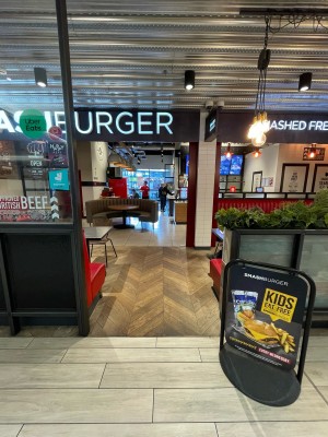 From inside shopping centre facing venue. Metal and glass divider on left. Divider with planters on right. Open entrance in between.