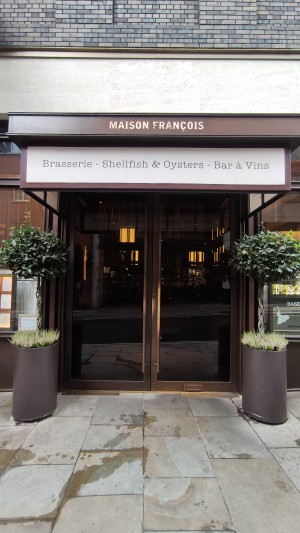 From sidewalk facing venue. Glass doors with brass frames. Push in centre via vertical brass handles to open. Planters on either side of doors.
