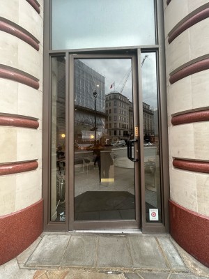 Single glass door with brown frame and small vertical black handle on right. Push to open.