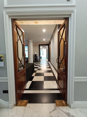 From back right of lobby, looking right. Glass doors with brown frame ahead. Brass handles in the middle. Push to open.