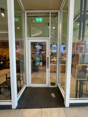 From inside shopping centre facing venue. Glass archway ahead with glass door in white frame. Pull vertical metal handle on right to open.