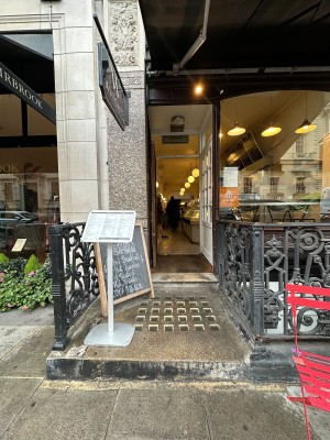 From street, facing venue. Step up before doorway ahead. White menu stand on left. A-frame board behind. Path ahead to doorway.