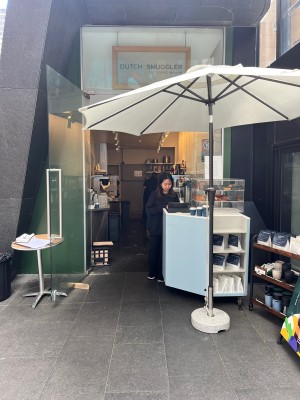 From sidewalk facing venue. Service counter ahead on right. Parasol and display shelf to its right. Table and glass door to staff area to its left.