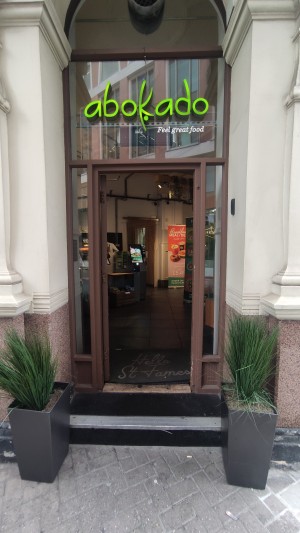 From sidewalk facing venue. Path ahead with planters on either side. One step up to glass door with brown frames. Push to right via metal handle.