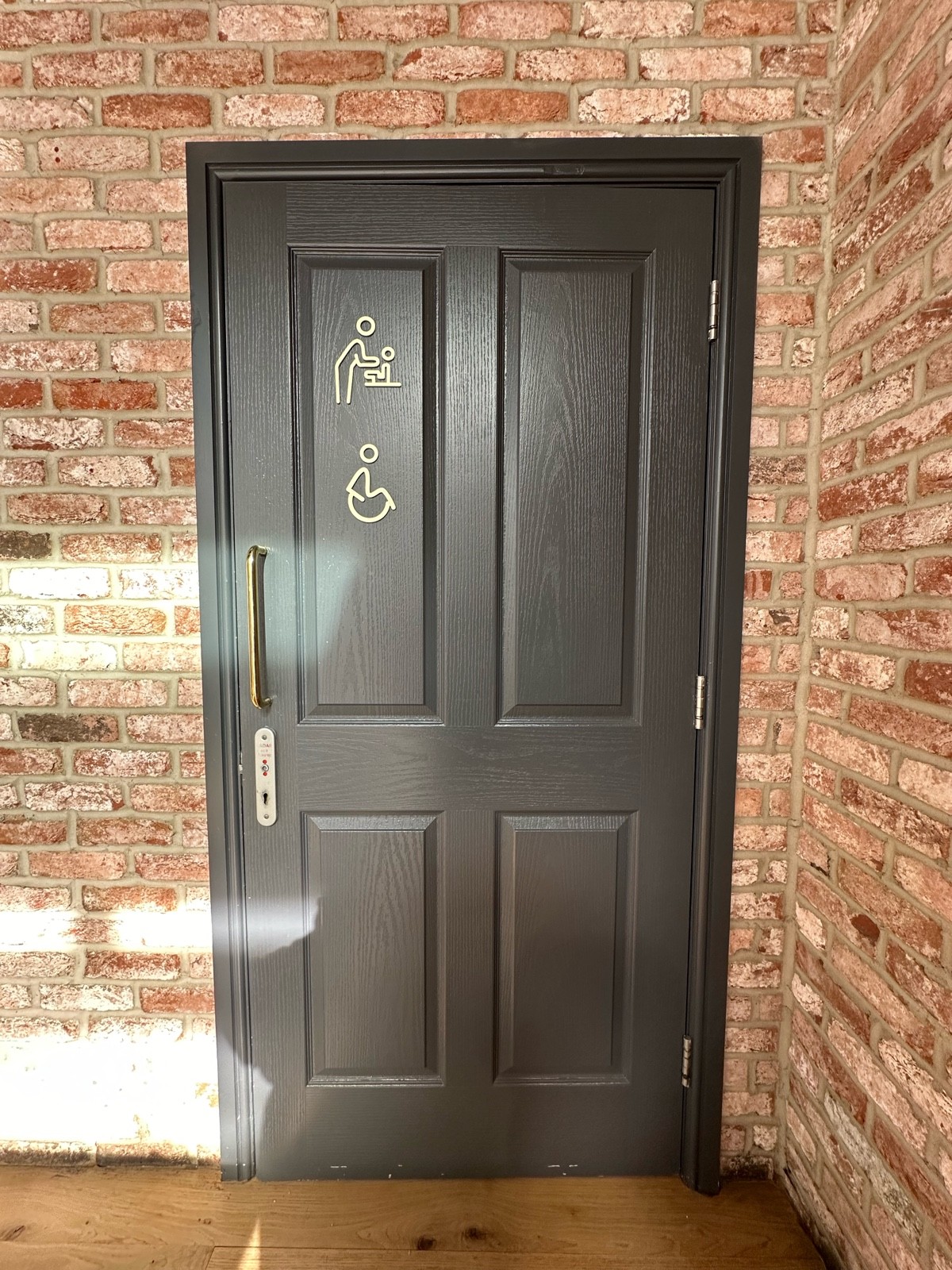 From end of half booths, facing left. Grey wooden bathroom door ahead. Gold metal vertical handle on left. Silver radar keyhole below it.