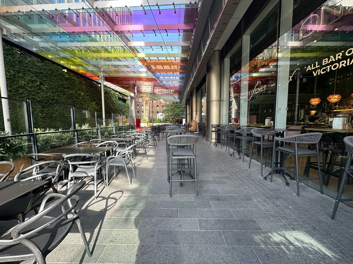 From opening, facing straight. Row of tables and chairs along left glass wall. High tables and stools ahead and along right. Path ahead to right.