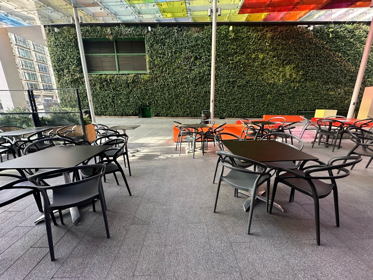 From bench, facing left. Two rows of tables and chairs ahead. Glass wall and planter ahead to left beyond seating.