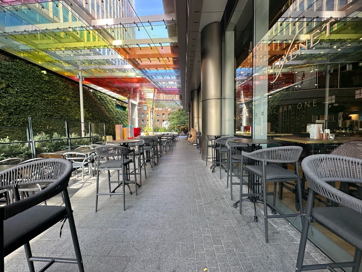 From start of high tables, facing left. High tables and stools along left and right of path ahead. Tables and chairs to back left.
