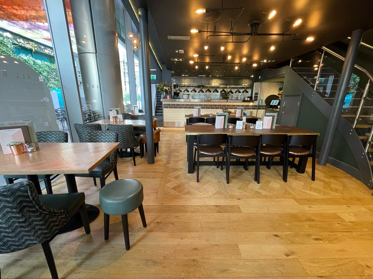 From after half booth, facing straight. Tables, chairs and stools along left. Path ahead. Large tables and chairs ahead to right. Stairs on right.