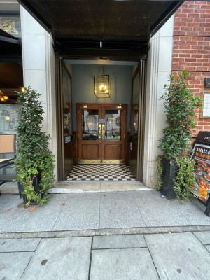 From pavement, facing straight. Path leads to wood-panelled glass doors. Push black handles in centre to open. Planters to left and right of doors.