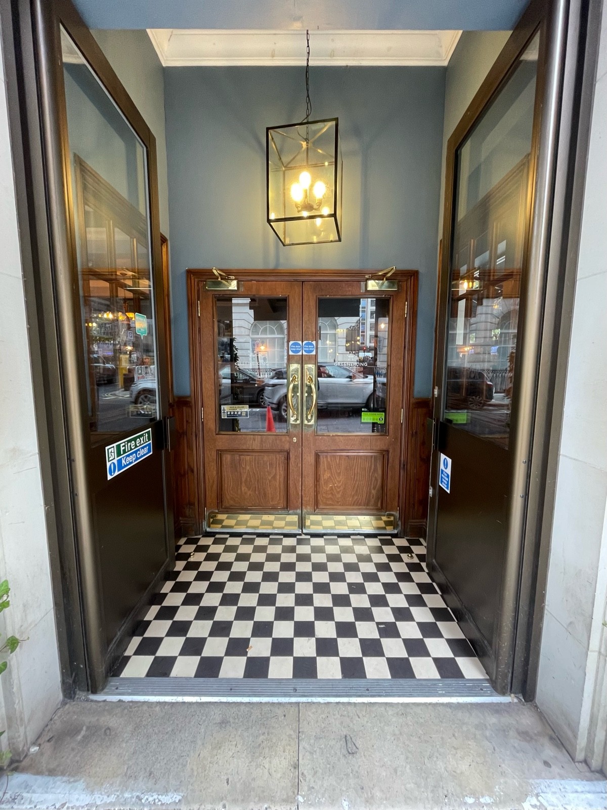 From doors, facing straight. Second set of wood-panelled glass doors ahead. Pull brass handles at centre open.