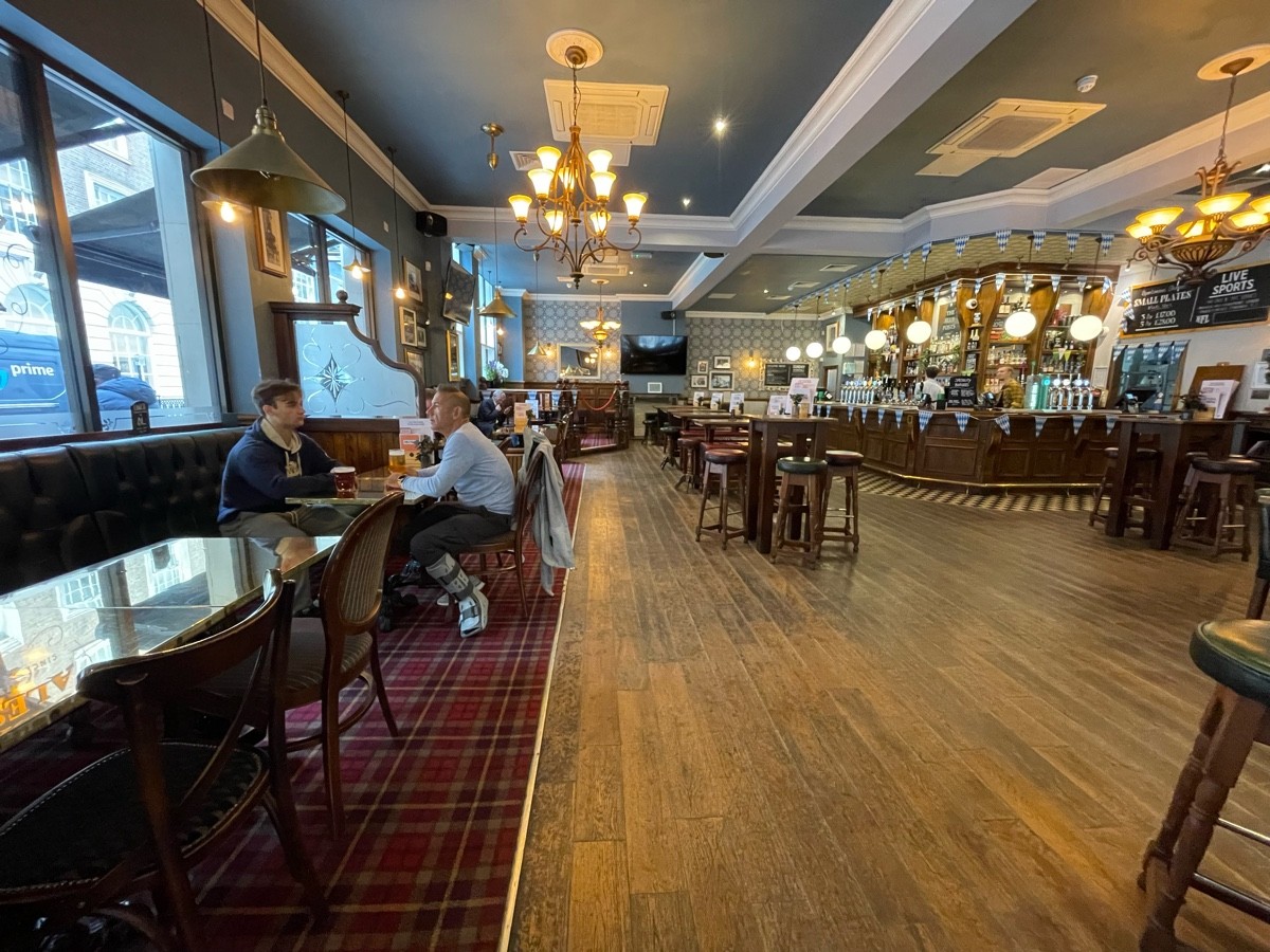 From entrance, facing left. Half booths with chairs along left. Path ahead. High tables and stools scattered ahead to right. Bar to back right.