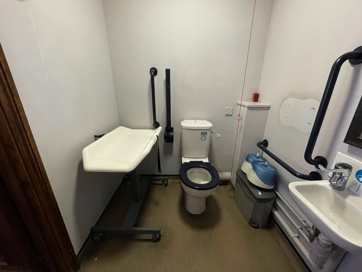 From door, facing left. Changing table on left. Toilet at back. Emergency cord on its right. Transfer space obstructed by table. Sink on right.