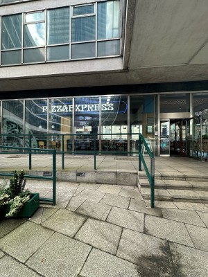 From pavement, facing straight. Opening to ramp ahead. Green railings to left and right of it. Steps ahead to right.